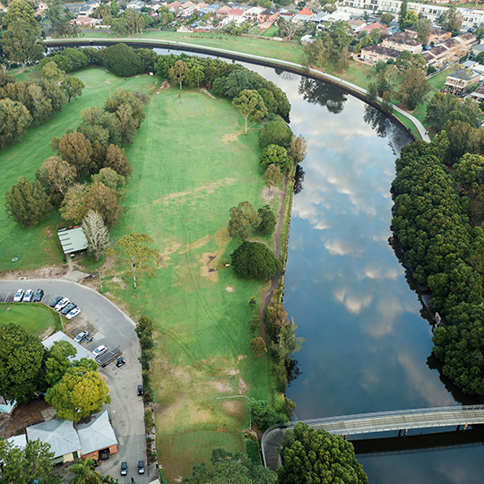 Aerial view of a golf course featuring the club house to the left, fairway centred and Cooks River to the right with the pedestrian bridge seen in the bottom right